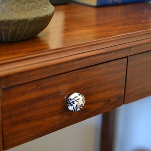 K&ouml;nnte beinhalten: Ein Holztisch mit zwei Schubladen und dekorativen Keramikkn&ouml;pfen. Die Schubladen sind aus dunklem Holz und haben eine glatte Oberfl&auml;che. Die Kn&ouml;pfe sind wei&szlig; mit schwarzen Blumenmustern.