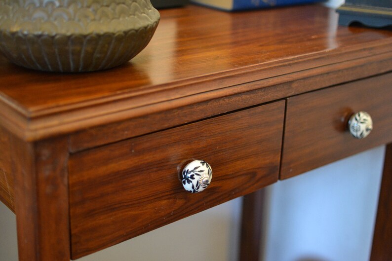 K&ouml;nnte beinhalten: Ein Holztisch mit zwei Schubladen und dekorativen Keramikkn&ouml;pfen. Die Schubladen sind aus dunklem Holz und haben eine glatte Oberfl&auml;che. Die Kn&ouml;pfe sind wei&szlig; mit schwarzen Blumenmustern.