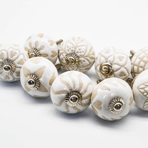 Peut inclure: Ensemble de boutons de meubles en céramique blanche avec divers motifs floraux et géométriques en relief. Chaque bouton présente une base métallique et un motif décoratif beige. Idéal pour la rénovation de meubles.