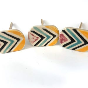 May include: Three ceramic cabinet knobs with a colourful chevron design. Each knob features a cream-coloured base with a pattern of black, teal, yellow, and burgundy chevrons. The knobs have silver-coloured stems.