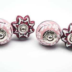 May include: Four ceramic cabinet knobs with floral designs. Two knobs are round with pink and white floral patterns and silver accents. Two knobs are star-shaped with red, white, and black floral patterns and silver accents.