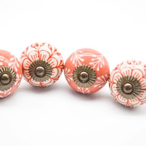 May include: Four ceramic drawer knobs with a white floral design on a coral orange background. Each knob has a brass colored screw in the center.