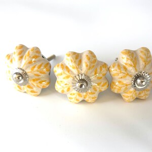 May include: Three ceramic flower-shaped cabinet knobs. The knobs are off-white with yellow leaf patterns and silver-coloured metal accents. Suitable for drawers and cabinets.
