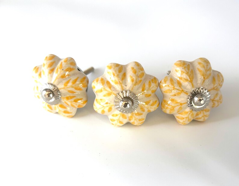 May include: Three ceramic flower-shaped cabinet knobs. The knobs are off-white with yellow leaf patterns and silver-coloured metal accents. Suitable for drawers and cabinets.