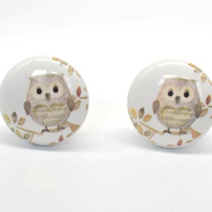 Op de afbeelding: Twee witte keramische kastknoppen met een geschilderd uilenontwerp. Elke knop heeft een bruine uil die op een tak zit met herfstkleurige bladeren. De knoppen zijn rond en hebben een glanzende afwerking.