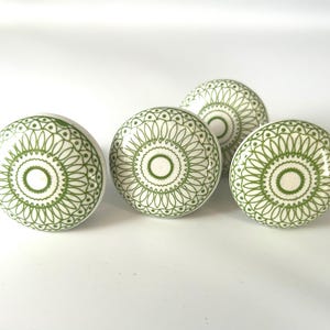 Op de afbeelding: Vier witte en groene decoratieve kastknoppen. Elke knop heeft een cirkelvormig ontwerp met een groen bloemmotief en een wit midden. De knoppen zijn rond en hebben een gladde, glanzende afwerking.