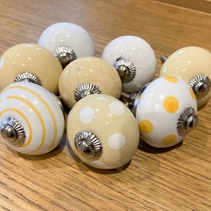 May include: Eight ceramic cabinet knobs in a variety of designs. The knobs are mainly white and cream, with patterns of stripes and polka dots in yellow and cream. Each knob has a silver metal base.