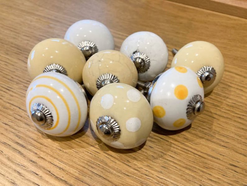 May include: Eight ceramic cabinet knobs in a variety of designs. The knobs are mainly white and cream, with patterns of stripes and polka dots in yellow and cream. Each knob has a silver metal base.