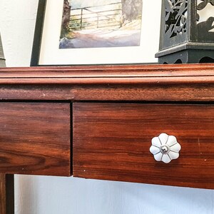 以下が含まれることがあります： 2 つの引き出しが付いた木製のコンソールテーブル。引き出しには、花柄の白いセラミックノブが付いています。テーブルは濃い茶色の仕上げです。