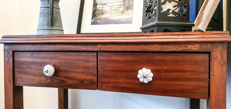 以下が含まれることがあります： 2 つの引き出しが付いた木製のコンソールテーブル。引き出しには、花柄の白いセラミックノブが付いています。テーブルは濃い茶色の仕上げです。