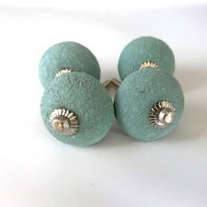 Puede incluir: Cuatro pomos de armario redondos, texturizados, de color verde azulado, con herrajes plateados. Los pomos est&aacute;n dispuestos en forma cuadrada, mostrando su dise&ntilde;o decorativo. Adecuados para a&ntilde;adir un toque de color y textura a los muebles.