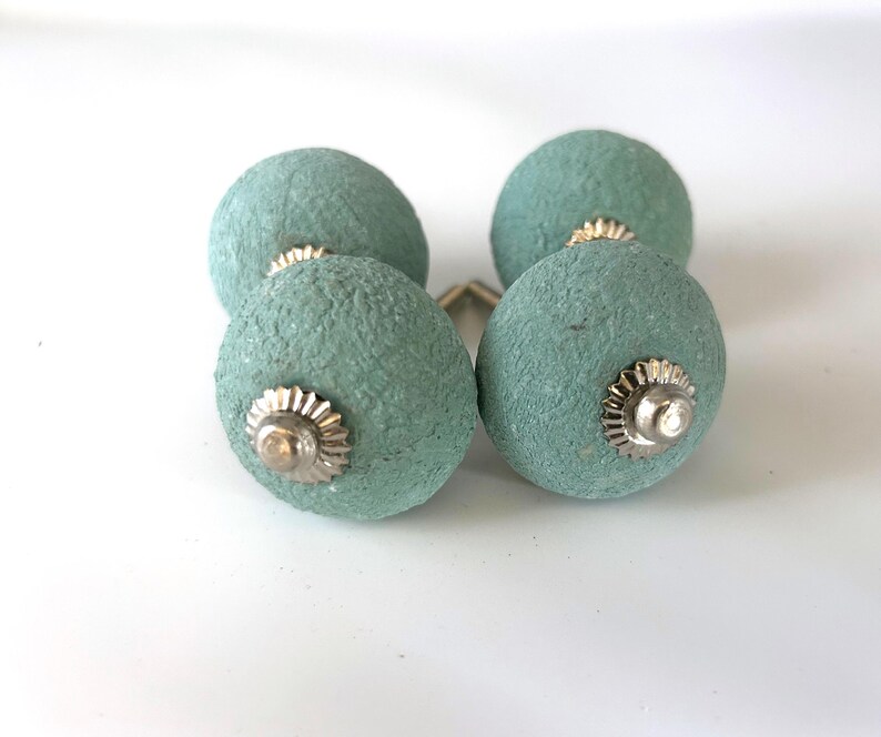 Puede incluir: Cuatro pomos de armario redondos, texturizados, de color verde azulado, con herrajes plateados. Los pomos est&aacute;n dispuestos en forma cuadrada, mostrando su dise&ntilde;o decorativo. Adecuados para a&ntilde;adir un toque de color y textura a los muebles.