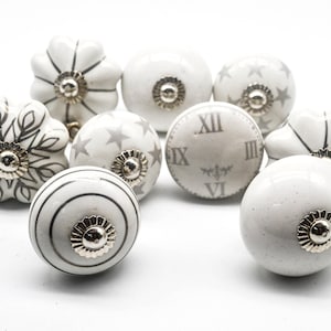 Op de afbeelding: Set van tien witte keramische kastknoppen met grijze bloemen-, sterren- en klokontwerpen. De knoppen hebben een zilveren metalen accent in het midden.