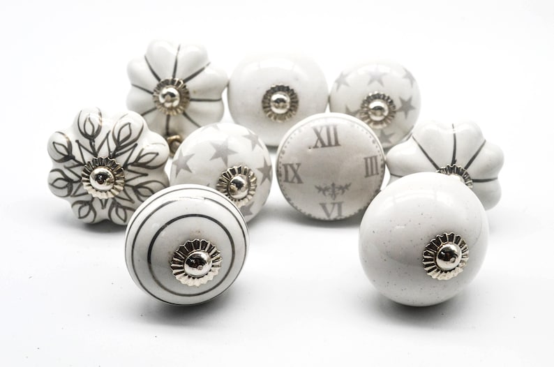 以下が含まれることがあります： グレーの花柄、星柄、時計柄の白いセラミック製のキャビネットノブ10個セット。ノブの中央にはシルバーの金属製のアクセントがあります。