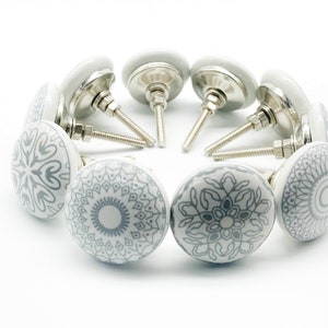 Può includere: Un set di dieci pomelli per armadi in ceramica con base in metallo argentato. I pomelli presentano un design a mandala grigio e bianco.
