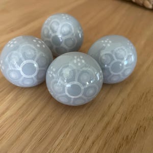 May include: Four pale grey decorative spheres with white floral patterns. The spheres are arranged on a wooden surface. Each sphere has a glossy finish and a unique design of circles and dots.