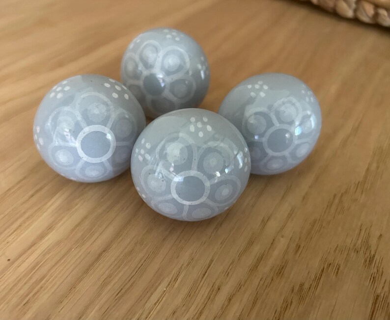May include: Four pale grey decorative spheres with white floral patterns. The spheres are arranged on a wooden surface. Each sphere has a glossy finish and a unique design of circles and dots.