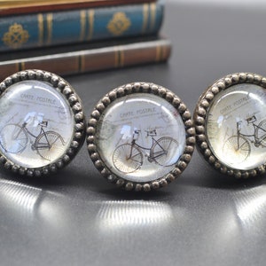 Peut inclure: Trois boutons de cabinet ton argent avec un dessus en verre transparent. Chaque bouton présente une image de carte postale vintage d'un vélo avec le texte "Carte Postale" imprimé sur la carte postale.