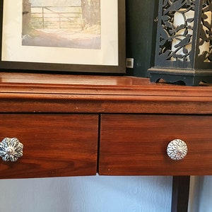 K&ouml;nnte beinhalten: Ein dunkler Holz-Beistelltisch mit zwei Schubladen. Die Schubladen haben dekorative silberne Kn&ouml;pfe. Der Tisch steht an einer wei&szlig;en Wand.