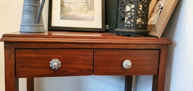 K&ouml;nnte beinhalten: Ein dunkler Holz-Beistelltisch mit zwei Schubladen. Die Schubladen haben dekorative silberne Kn&ouml;pfe. Der Tisch steht an einer wei&szlig;en Wand.