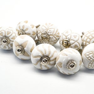 Op de afbeelding: Een set van tien witte keramische kastknoppen met een beige bloemenmotief. Elke knop heeft een gouden metalen basis.