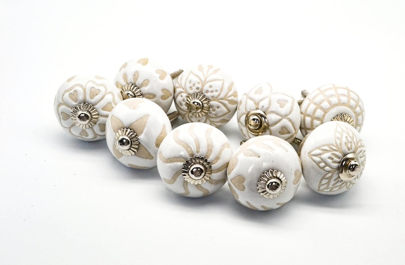 Op de afbeelding: Een set van tien witte keramische kastknoppen met een beige bloemenmotief. Elke knop heeft een gouden metalen basis.