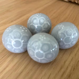 May include: Four light gray decorative spheres with white patterned designs. The spheres are arranged on a wooden surface. The spheres have a glossy finish and a detailed, repeating pattern.