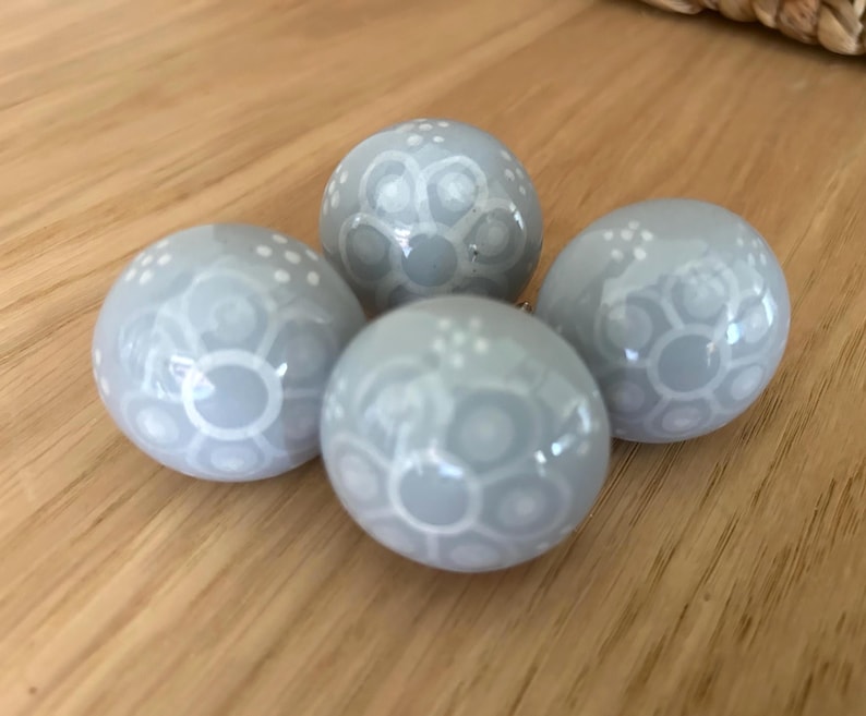 May include: Four pale grey decorative spheres with white patterned designs. The spheres are arranged on a wooden surface. The spheres have a glossy finish and a detailed, repeating pattern.