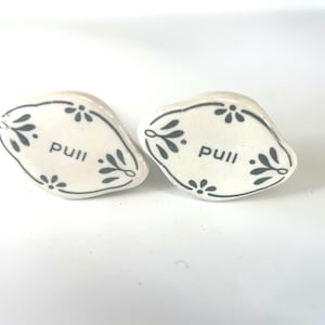Peut inclure: Deux boutons de meuble en céramique blanche avec un motif floral et le mot "pull" en noir. Les boutons ont une forme légèrement irrégulière et conviennent aux tiroirs ou aux armoires.