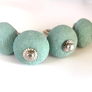 Puede incluir: Cuatro pomos de armario de color aguamarina con un acabado texturizado. Cada pomo tiene una roseta decorativa plateada y un tornillo plateado. Los pomos son redondos y parecen estar hechos de un material compuesto.