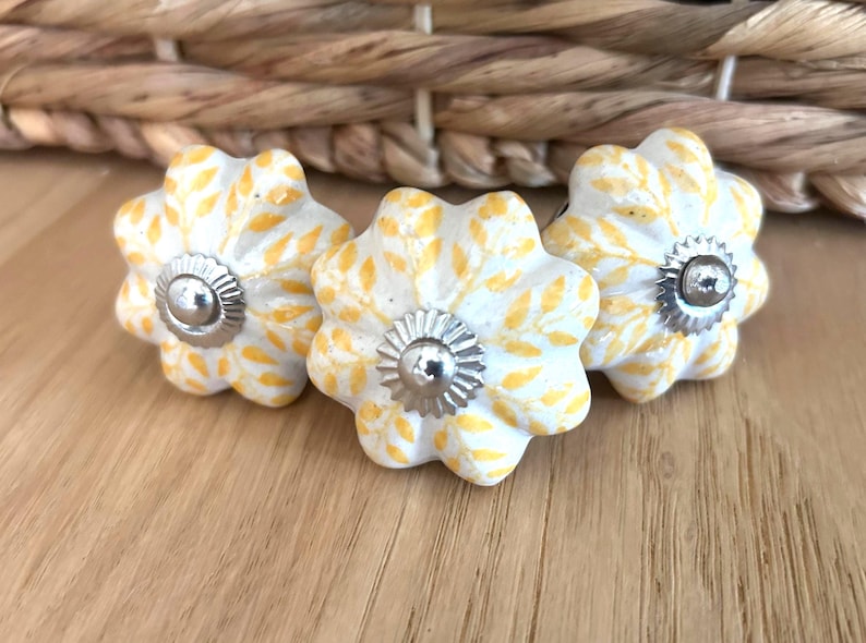 May include: Three flower-shaped cabinet knobs. Each knob has a white base with yellow leaf patterns and a silver metal centre. The knobs are arranged on a light wood surface.