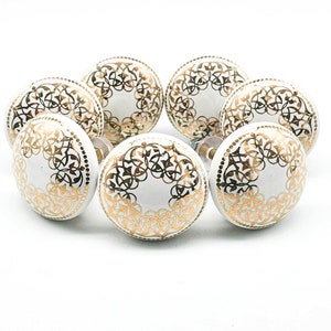 May include: A collection of eight white ceramic cabinet knobs with a gold filigree design. The knobs are arranged in a circular pattern against a white background, showcasing their decorative detail. Each knob has a diameter of approximately 1.5 inches.