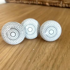 Peut inclure: Trois boutons de meuble en céramique blanche et grise avec un motif mandala décoratif. Les boutons sont ronds et présentent un cercle central entouré d'un motif détaillé. Ils sont disposés sur une surface en bois clair.