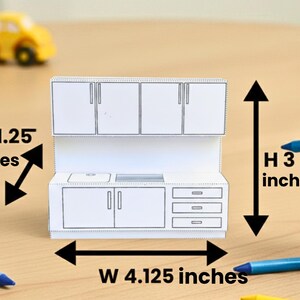 A papercraft miniature kitchen counter and upper cabinets with dimension arrows and measurement text.