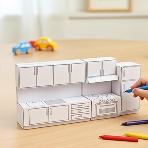A child's hand colors a paper craft miniature kitchen, featuring a sink, stove, and refrigerator, with toy cars in the background.