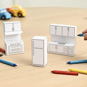A child's hand holds a blue crayon near three uncolored paper craft miniature kitchen pieces: a stove, a refrigerator, and a sink unit.