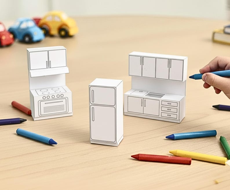 A child's hand holds a blue crayon near three uncolored paper craft miniature kitchen pieces: a stove, a refrigerator, and a sink unit.