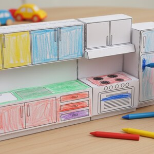 A child's hand colors a paper craft miniature kitchen, with sections already colored red, blue, green, and yellow, and crayons scattered around.