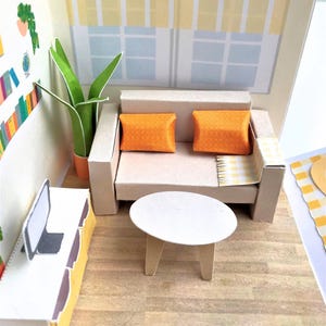 Puede incluir: Una escena de sala de estar en miniatura con un sofá beige, dos cojines naranjas y una pequeña mesa blanca. Una planta verde alta en una maceta naranja está al lado del sofá. La habitación tiene una ventana con una cortina a rayas amarillas y blancas.