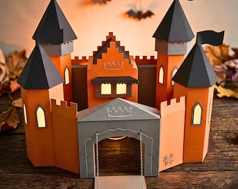 Puede incluir: Un castillo de papel con paredes naranjas, techos cónicos grises con banderas negras y una puerta gris con puente levadizo. La estructura incluye ventanas iluminadas, un contorno de corona en la puerta y una pequeña araña recortada en el muro.