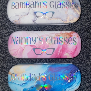 Pode incluir: Três estojos para óculos com padrões de mármore diferentes, com os nomes "BamBam's Glasses", "Nanny's Glasses" e "Grandad's Glasses" impressos em glitter prateado.