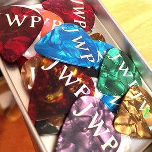 May include: A tin container filled with a variety of colorful guitar picks. The picks are made of different materials and have the initials "JWP" printed on them.