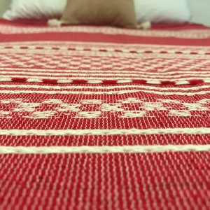 May include: A woven textile blanket featuring a vibrant red base color with intricate white geometric patterns and horizontal stripes. The fabric displays a textured, interlaced weave construction that covers the surface in a repeating design.