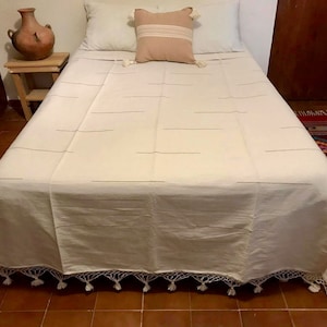 May include: A white bedspread with a subtle striped pattern and a white crocheted trim with tassels. The bedspread is on a bed with white pillows and a beige throw pillow with an embroidered design.