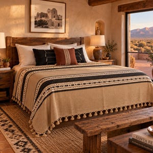 May include: A beige bedspread with black horizontal stripes and a repeating geometric pattern, finished with a fringe of small tassels along the bottom edge. The textile is draped over a bed, featuring a textured weave and neutral color palette.