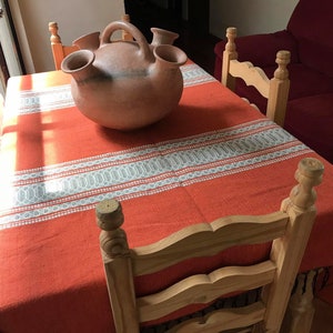 May include: A terracotta pitcher with multiple spouts sits on a red and white striped tablecloth. The tablecloth is on a wooden table with four wooden chairs.