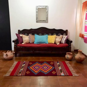 May include: A dark wood couch with a red cushion and colorful pillows. The couch is in front of a white wall with a silver framed mirror above it. A colorful rug with fringe is on the floor in front of the couch. Two terracotta pots are on the floor on either side of the rug.