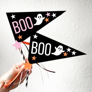 Op de afbeelding: Twee zwarte driehoekige vlaggen met het woord "BOO" in witte letters en een witte geest op elke vlag. De vlaggen zijn bevestigd aan oranje-wit gestreepte rietjes met roze en oranje sterren.
