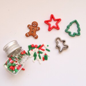 May include: A glass jar filled with red, green, and white sprinkles.  A gingerbread man cookie cutter, a star cookie cutter, a Christmas tree cookie cutter, and a person cookie cutter are scattered around the jar.
