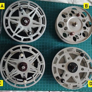 Könnte beinhalten: Vier Angelrollen im Vintage-Stil mit silbernen Metallrahmen und dunklen Zentren sind auf einer grünen Schneidematte angeordnet. Die Rollen haben verschiedene Speichendesigns und kreisförmige Öffnungen. Jede Rolle hat einen Griff und eine zentrale Nabe.